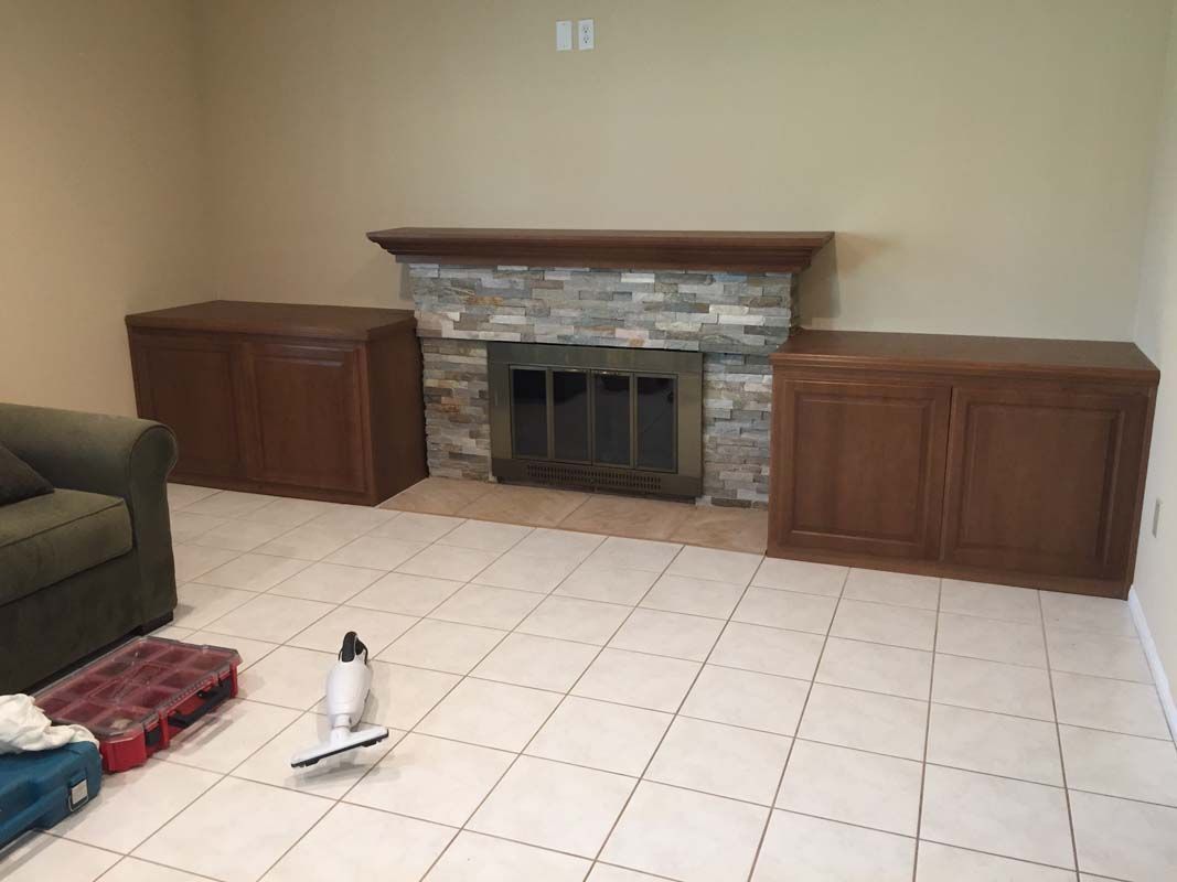 Fireplace with stone surround, flanked by two wooden cabinets, in a room with tile flooring.