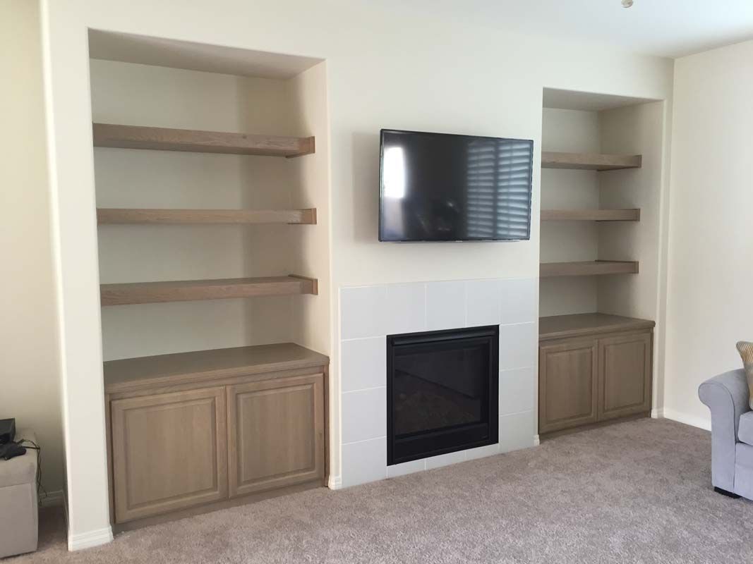 Built-in shelves and cabinets flank a fireplace and television in a living room with beige walls and carpet.