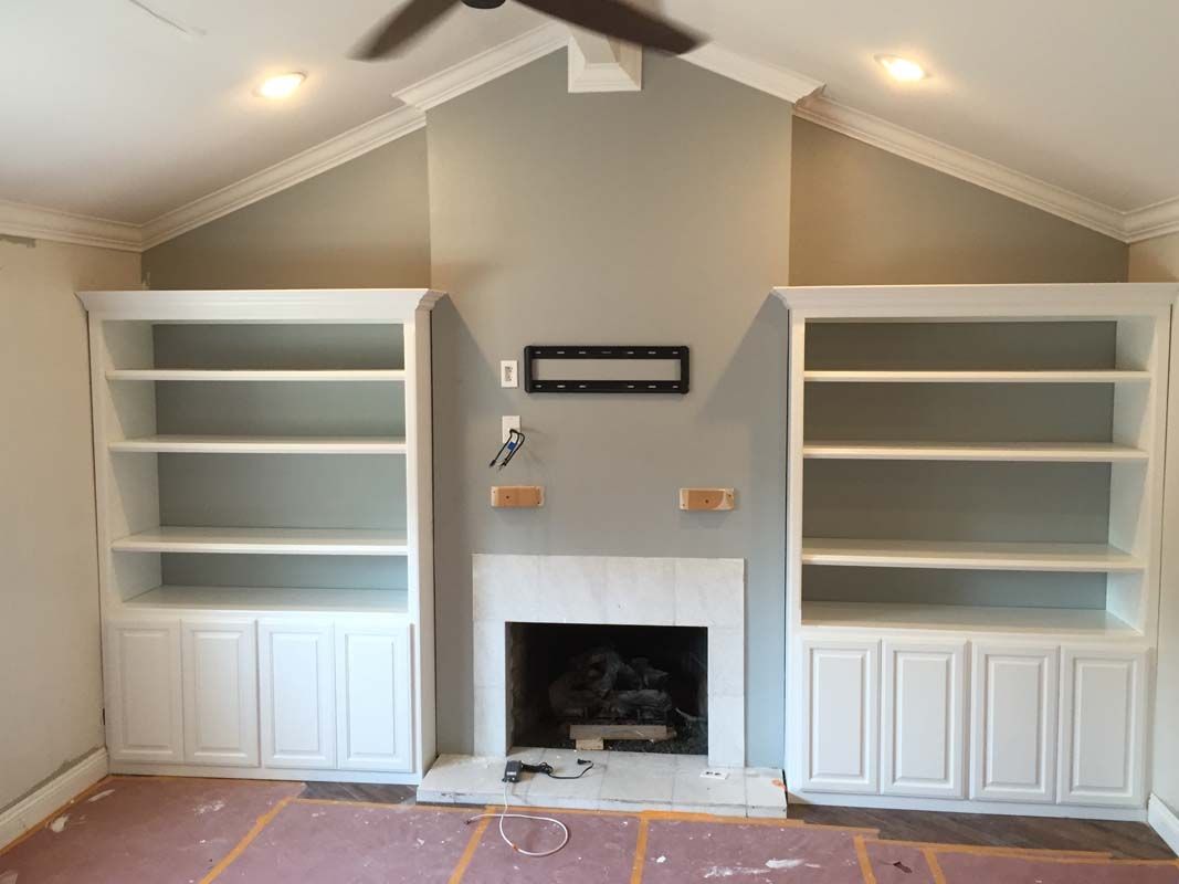 Built-in white bookshelves flank a fireplace, with gray wall, unfinished flooring, and ceiling fan.