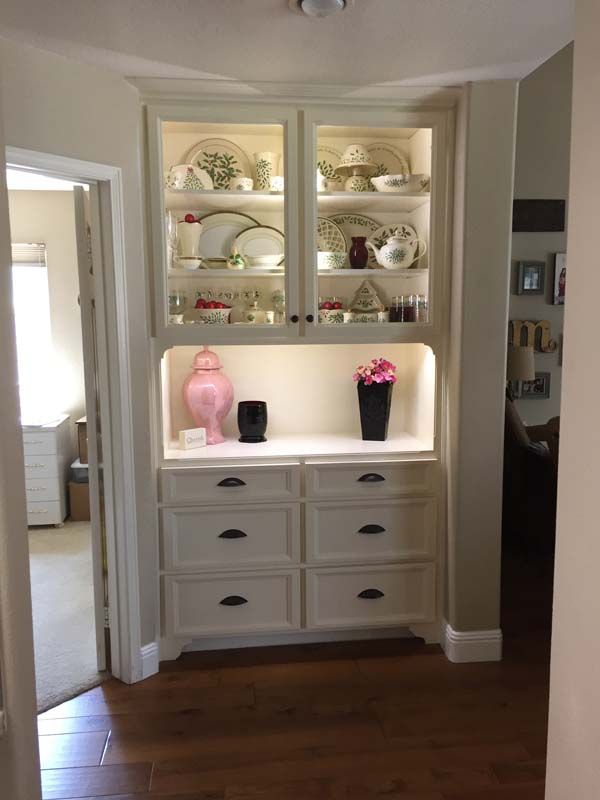 Built-in white cabinet in hallway with glass doors, illuminated shelves holding dishes. Drawers below.