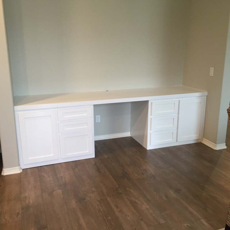 White built-in desk with cabinets and drawers, set against a light-colored wall with wood flooring.