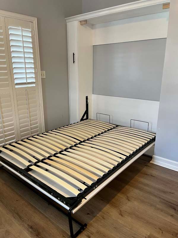 A white and wooden wall bed frame is lowered from its cabinet against a gray wall.