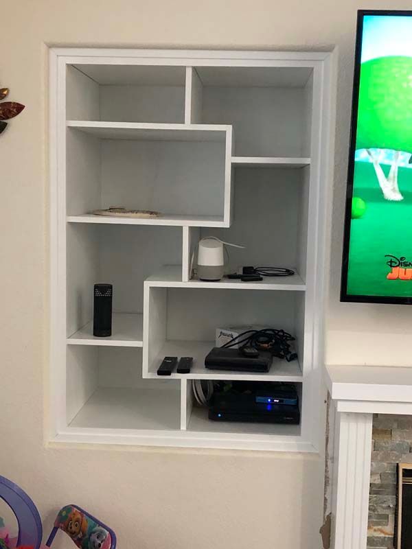 White built-in shelving unit with a stepped design, holding electronics, a smart speaker, and decorative items.