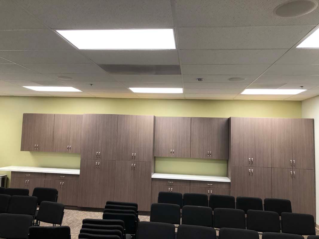 Conference room with rows of black chairs facing cabinetry. Beige walls, white ceiling lights.