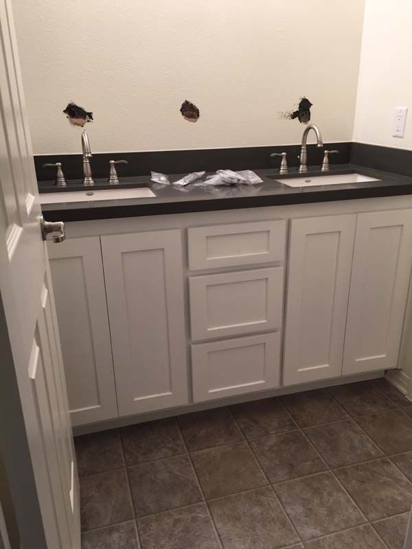 White bathroom vanity with two sinks, black countertop, and three drawers. Brown tiled floor.