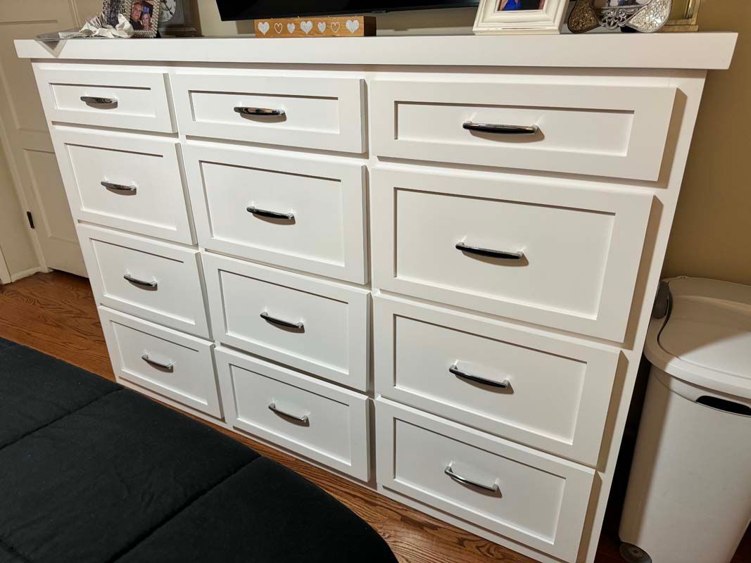 White dresser with 12 drawers, black hardware. Wooden floor, wall, and black bed are also visible.
