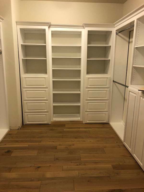 Built-in white shelves and drawers in a closet with wood flooring and hanging rod.