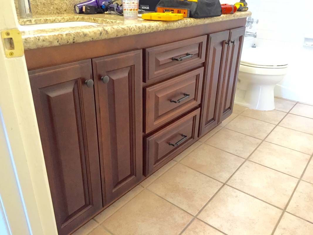 Brown bathroom vanity with drawers and doors, tile floor, and a toilet in the background.