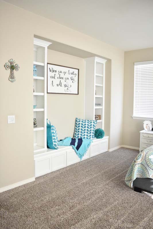 Built-in bedroom reading nook with white bookshelves, blue pillows, and blanket; neutral walls and carpet.