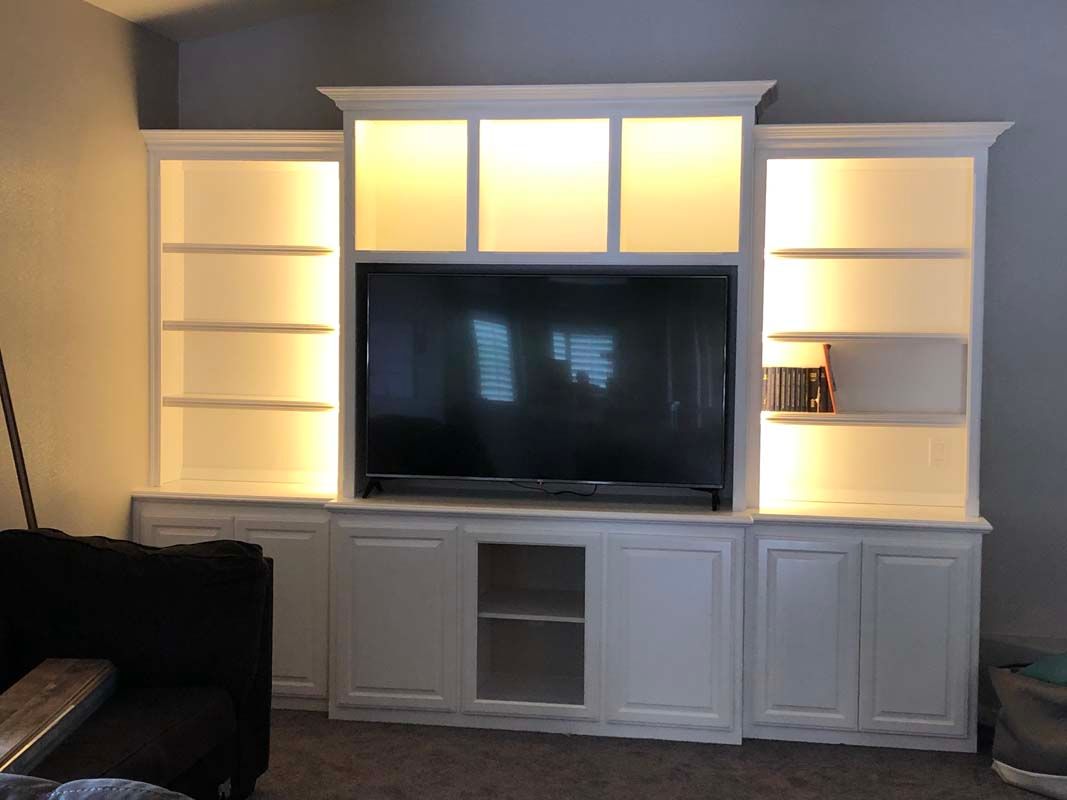 White built-in entertainment center with shelving, cabinets, and a television. Lit with warm lights.