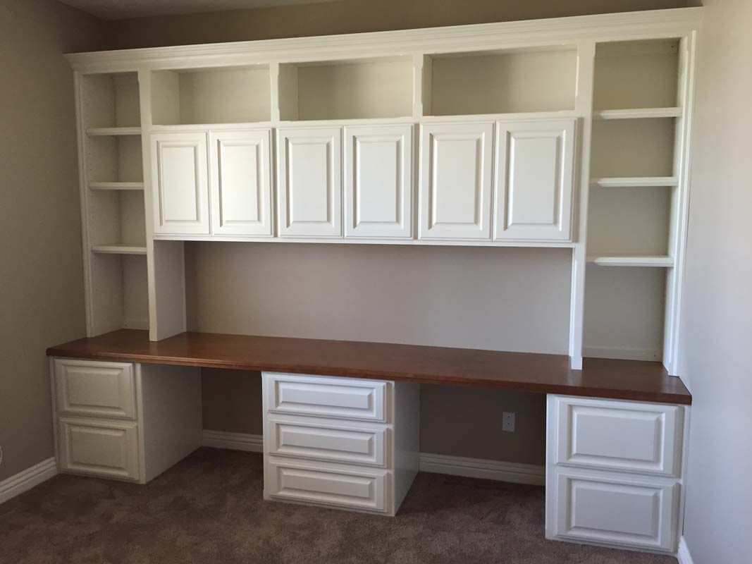 Built-in white desk with cabinets, shelves, and a wooden countertop against a neutral wall; room with carpet.