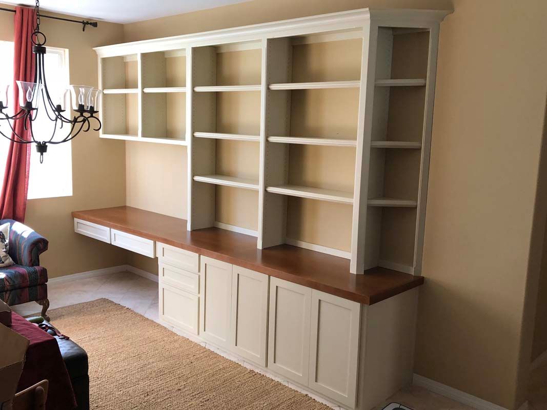 Built-in cream-colored bookcase and desk with a dark wood countertop. Shelves are above and cabinets below.