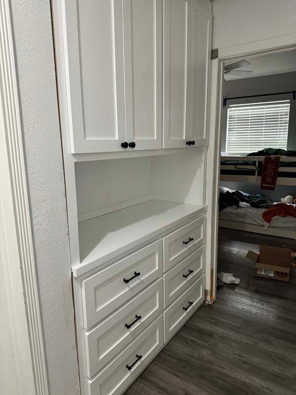 White built-in storage unit with cabinets, open shelf, and drawers in a hallway, leading to a cluttered bedroom.