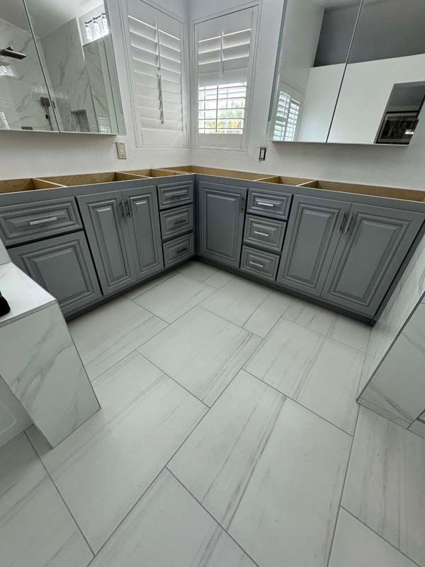 Gray bathroom cabinets, drawers, and white tiled floor under mirrors and window with shutters.
