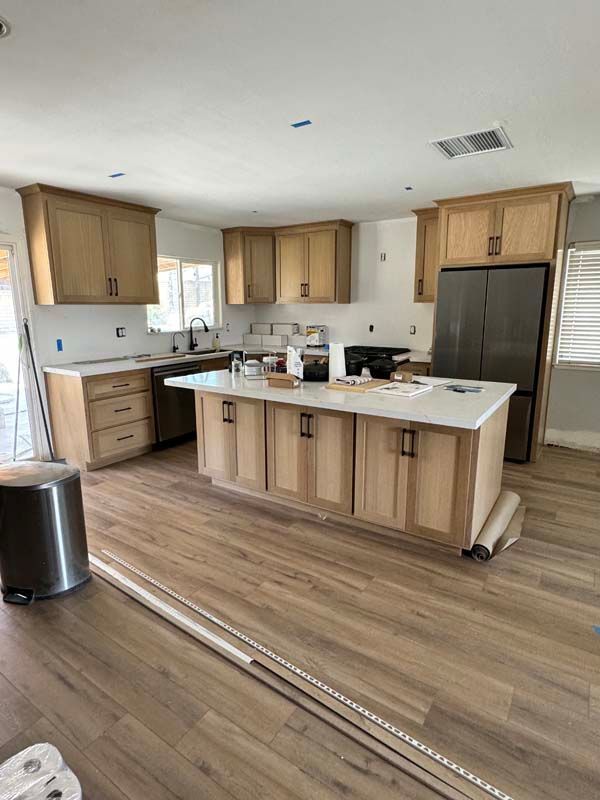 Kitchen remodel: Light wood cabinets, white countertops, island, stainless steel refrigerator, and vinyl wood-look flooring.