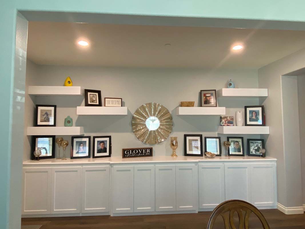 White built-in shelving and cabinets with framed photos, decor, and a round gold mirror on a light blue wall.