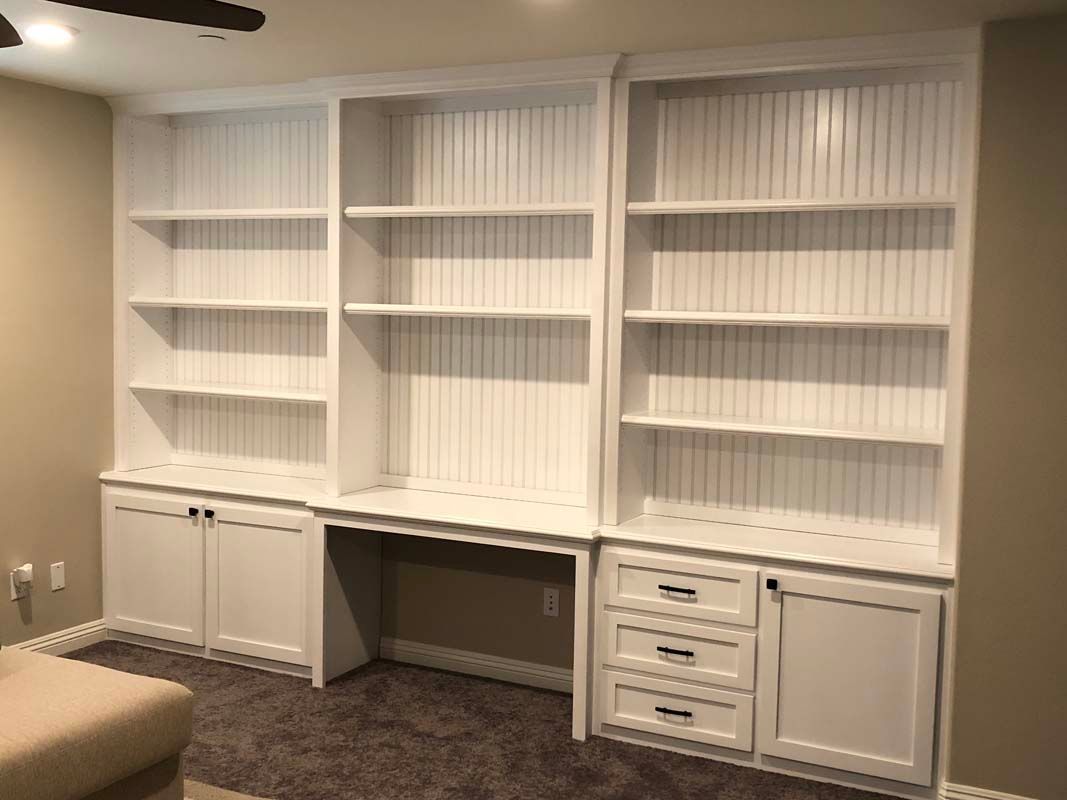 White built-in bookshelves with a desk area, cabinet doors, and drawers. Beige wall and dark carpet.