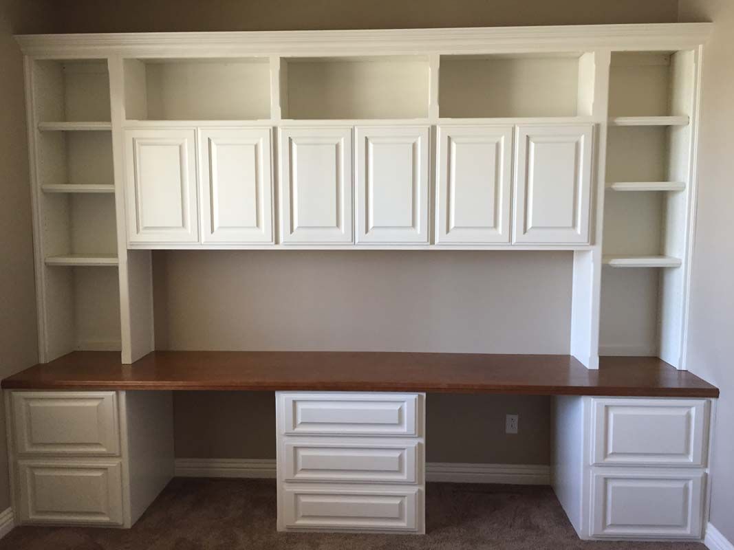 Built-in white desk with cabinets, shelves, and a wooden countertop against a beige wall.