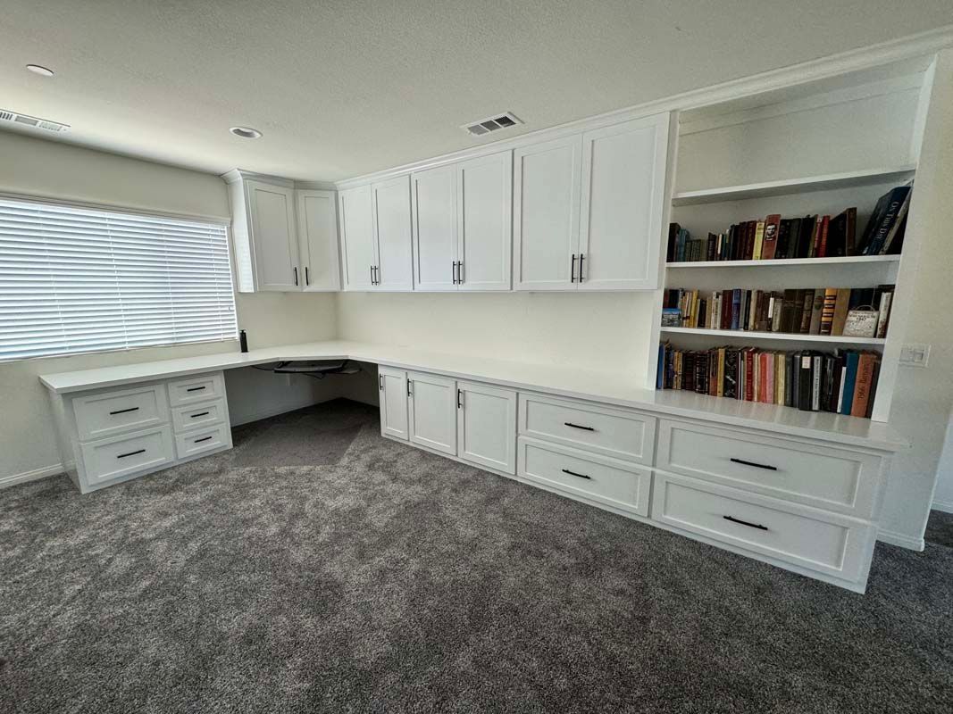 White built-in office furniture in a room. Includes desk, cabinets, shelves, and books on a gray carpet.