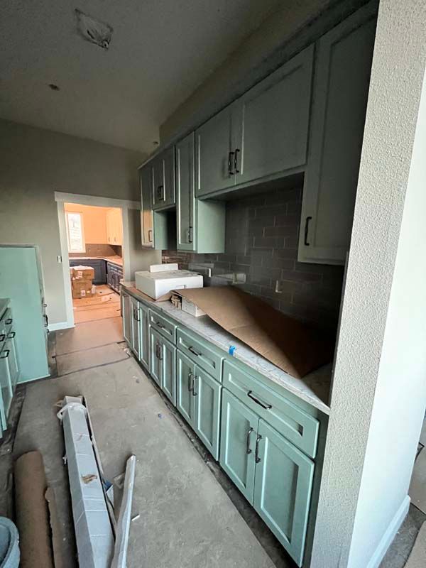 Kitchen under construction; light green cabinets, gray countertop, backsplash, doorway to another room.