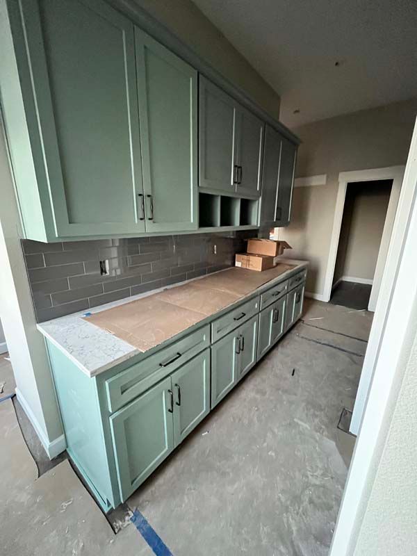 Blue-green kitchen cabinets with gray tile backsplash and light-colored countertop under construction.
