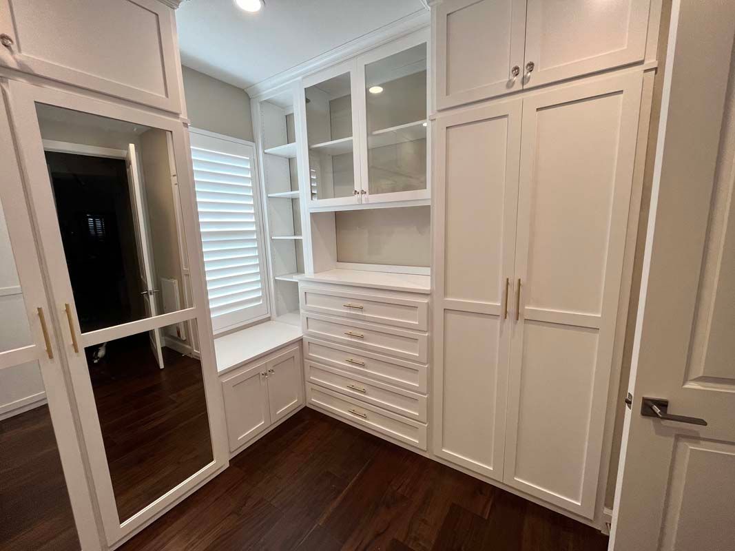White walk-in closet with built-in cabinetry, drawers, window seat, mirror, and dark wood flooring.