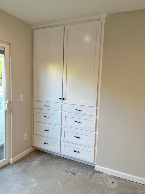 White built-in storage cabinet with doors and drawers against a beige wall. Next to a sliding glass door.