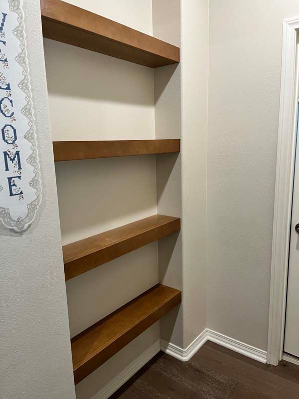 Four wooden shelves built into a wall niche, painted beige. A welcome sign hangs on the left.