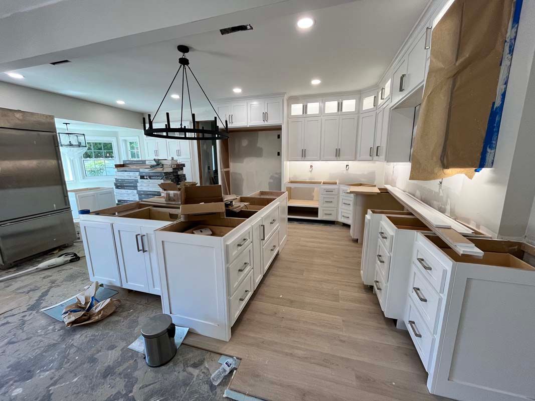 Kitchen cabinets under construction, with an island and curved countertop area. White cabinets, light wood floor, and exposed walls.
