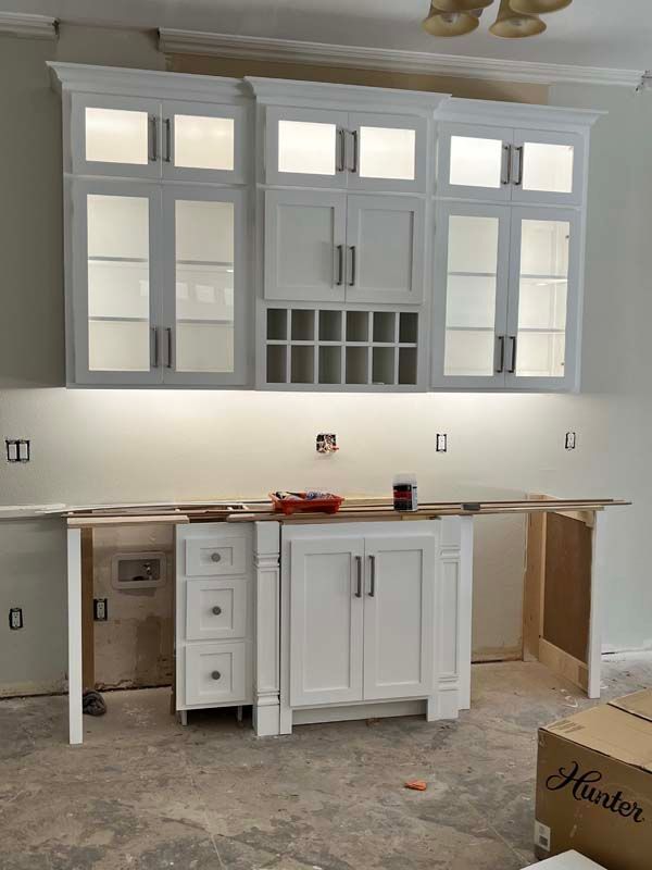 White kitchen cabinets under construction. Upper cabinets with glass fronts, lower cabinet with drawers and doors, and unfinished countertop.
