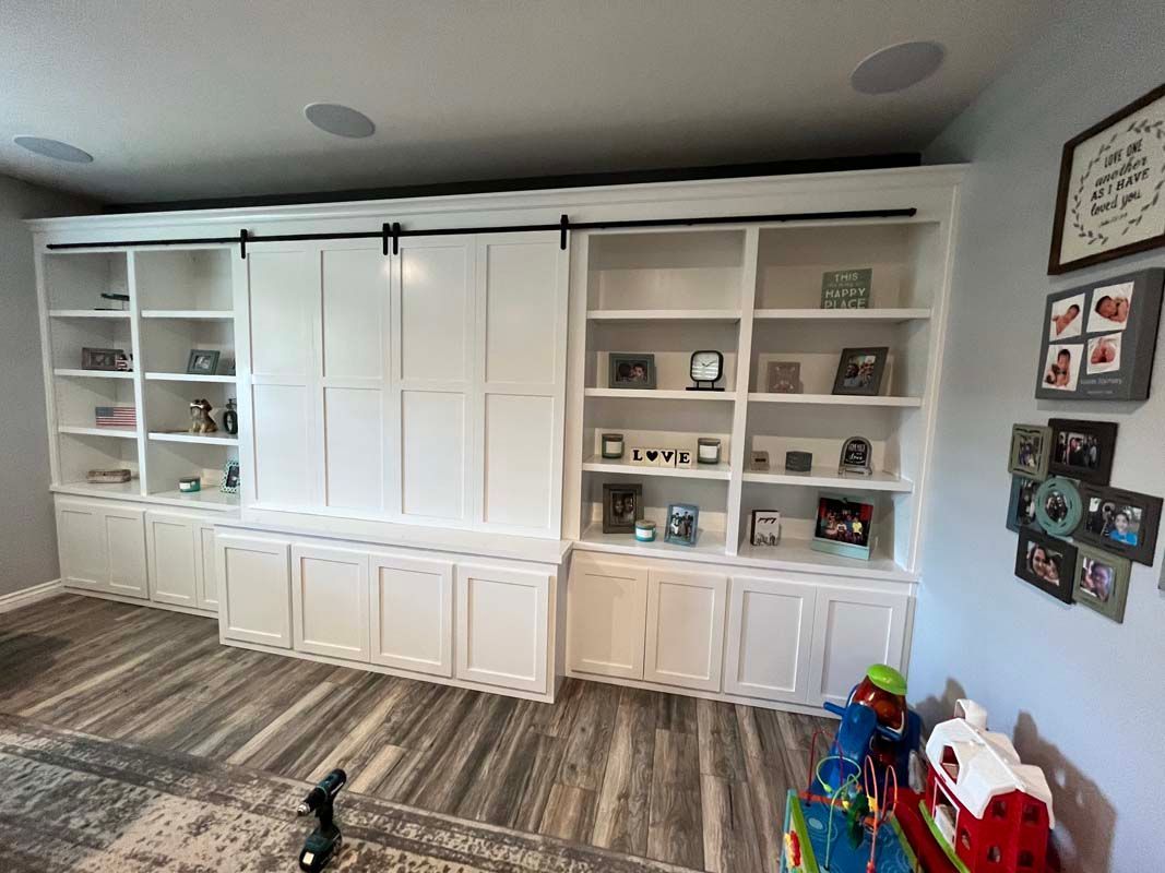 White built-in shelving with cabinets and sliding doors; gray flooring, toys on floor, pictures on wall.