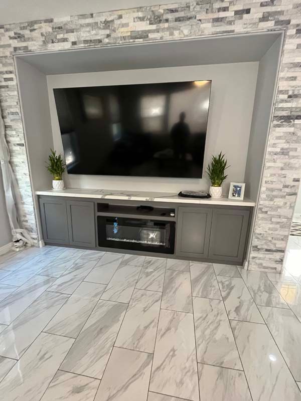 Built-in gray entertainment center with fireplace, TV, and cabinets, framed by tile, in a room with marble-look flooring.