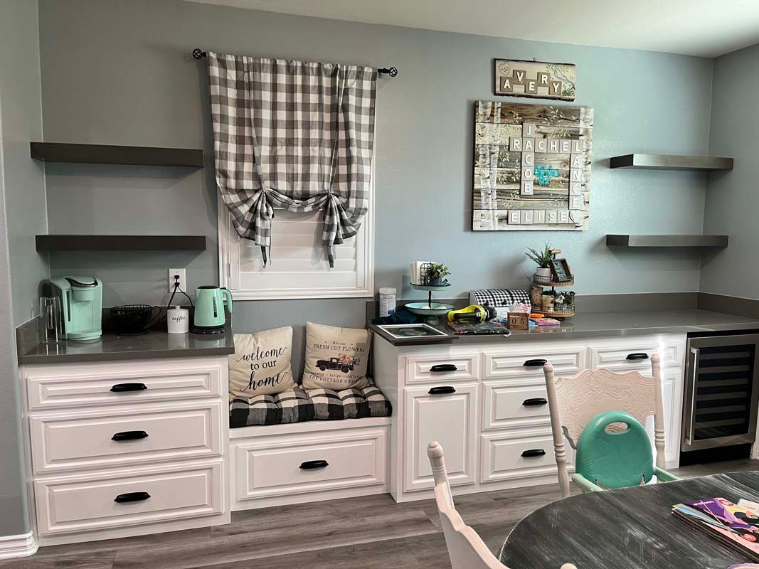 A built-in kitchen area with white cabinets, gray countertops, and open shelving. There is a window with a plaid curtain and a seating bench.