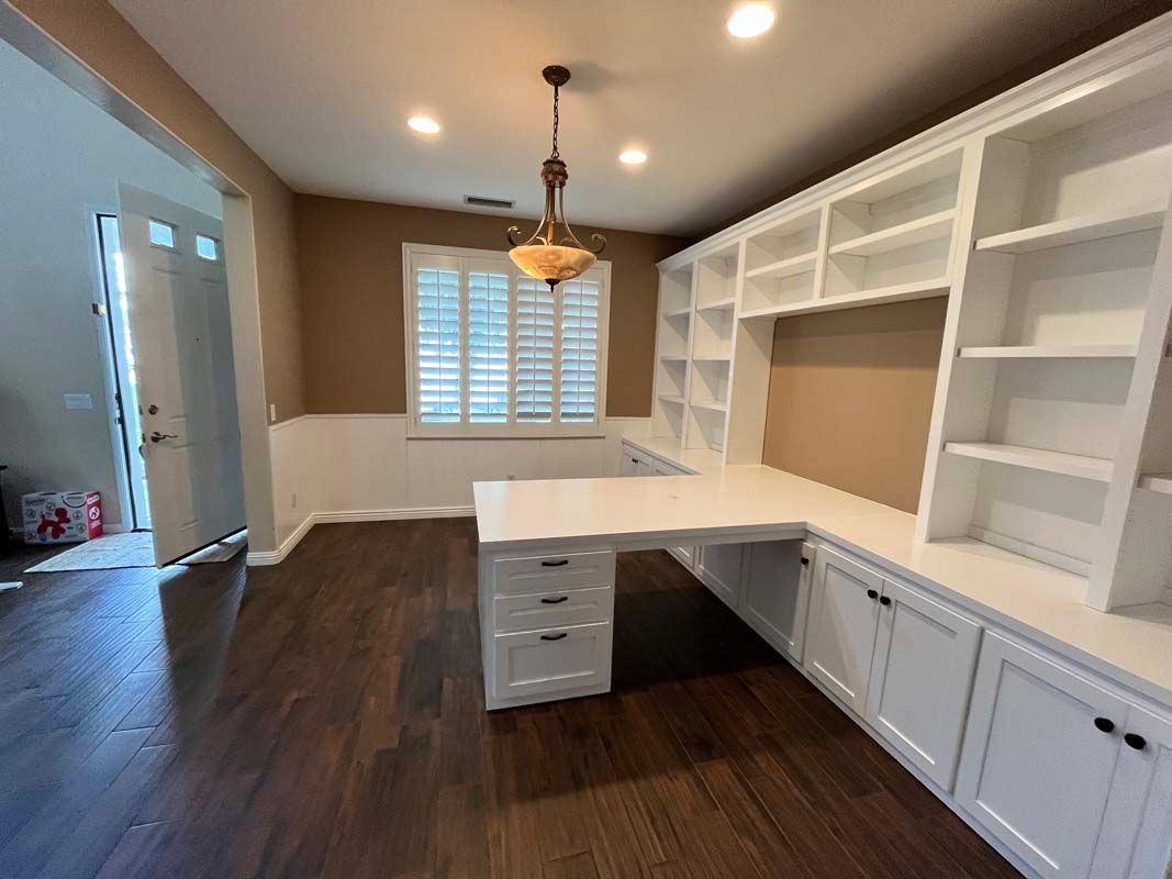 Home office with white built-in desk, shelving, dark wood floors, and shutters.