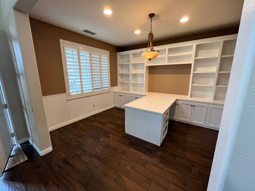 Office with built-in white shelves, desk, and brown wooden floors. Brown walls, window with shutters.