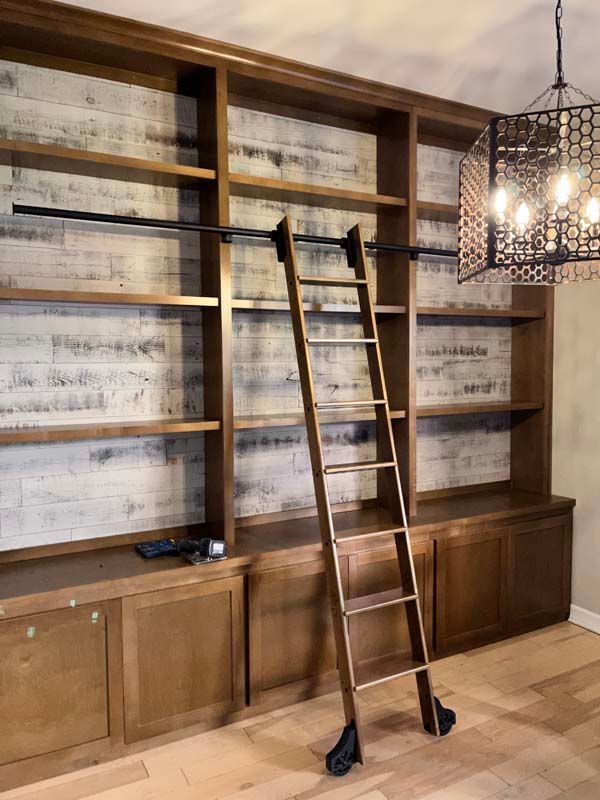 Wooden library with ladder, shelves, and cabinets. Black metal track and ornate chandelier.