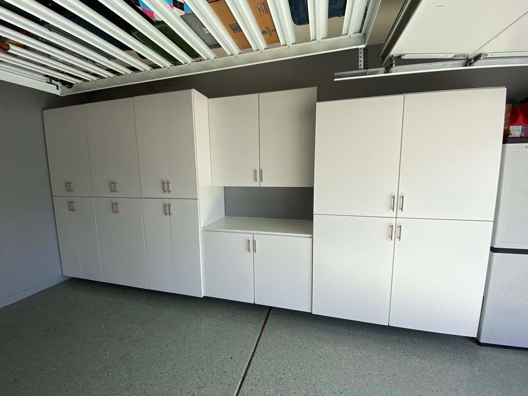 White storage cabinets in a garage. Cabinets include tall, short, and wall-mounted units. Garage floor is gray.