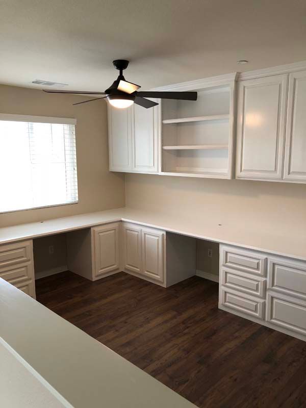 White cabinets and desks in a home office with a dark ceiling fan and window with blinds.