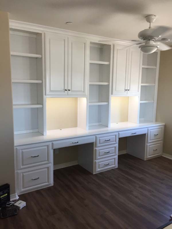 White built-in desk unit with cabinets, shelves, and recessed lighting against a light-colored wall. Dark wood-look floor.