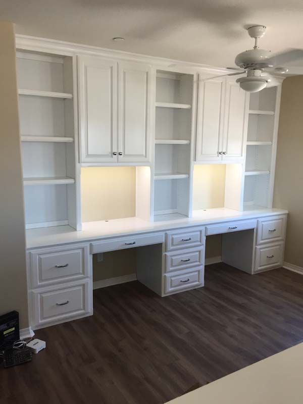 Built-in white cabinets with desks and shelving, under-cabinet lighting, set in a room with wood-look flooring.