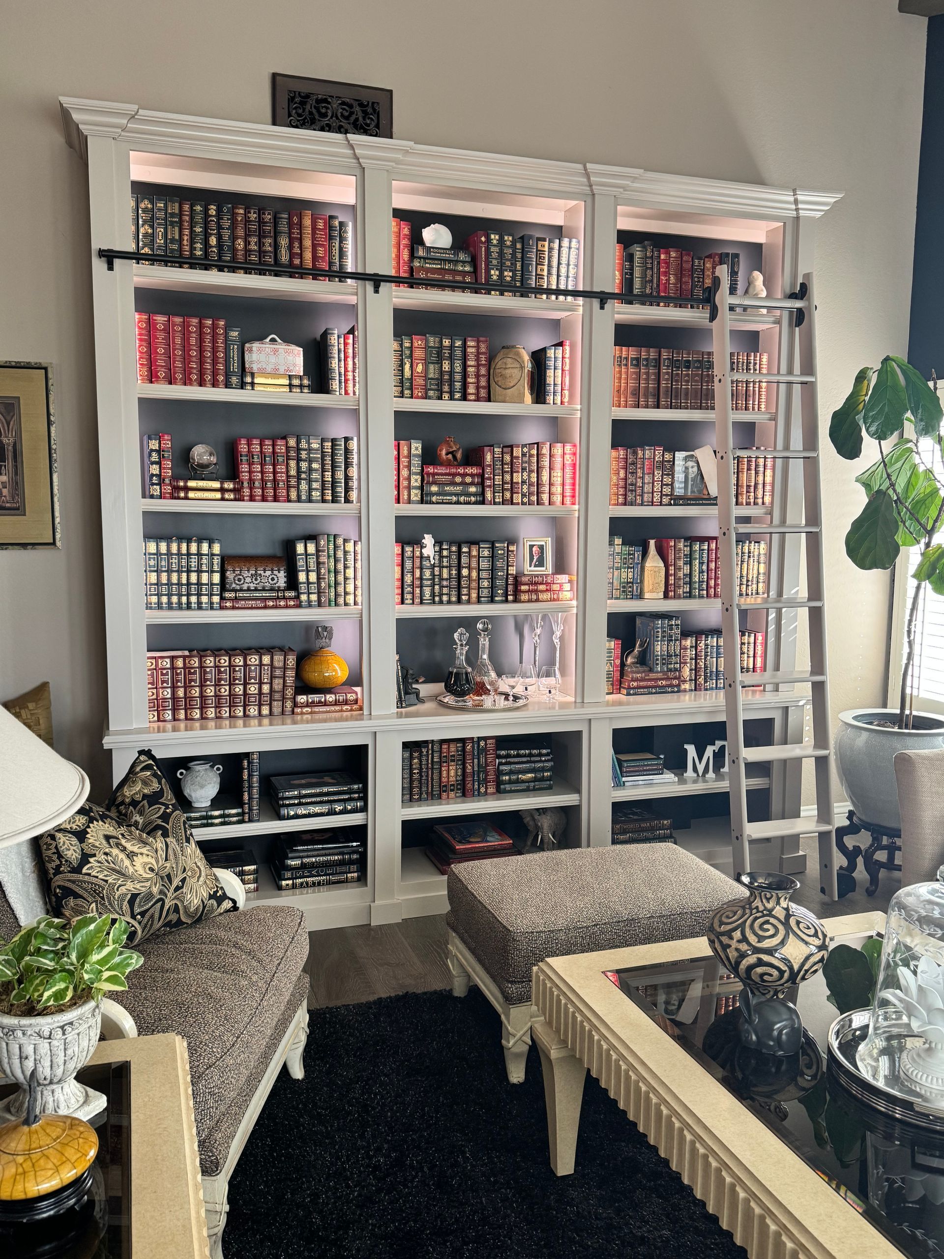 White built-in bookshelves with books, decorative items, and a rolling ladder. Living room setting with chairs and a coffee table.