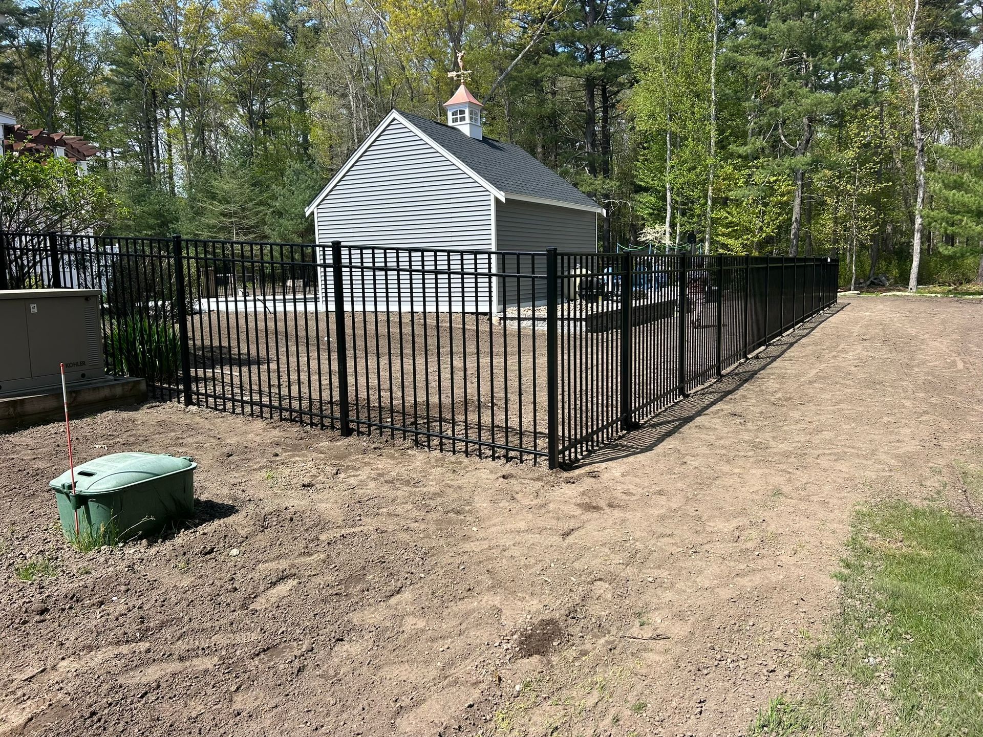 Black metal fence around a small gray shed beside a dirt path in a wooded area