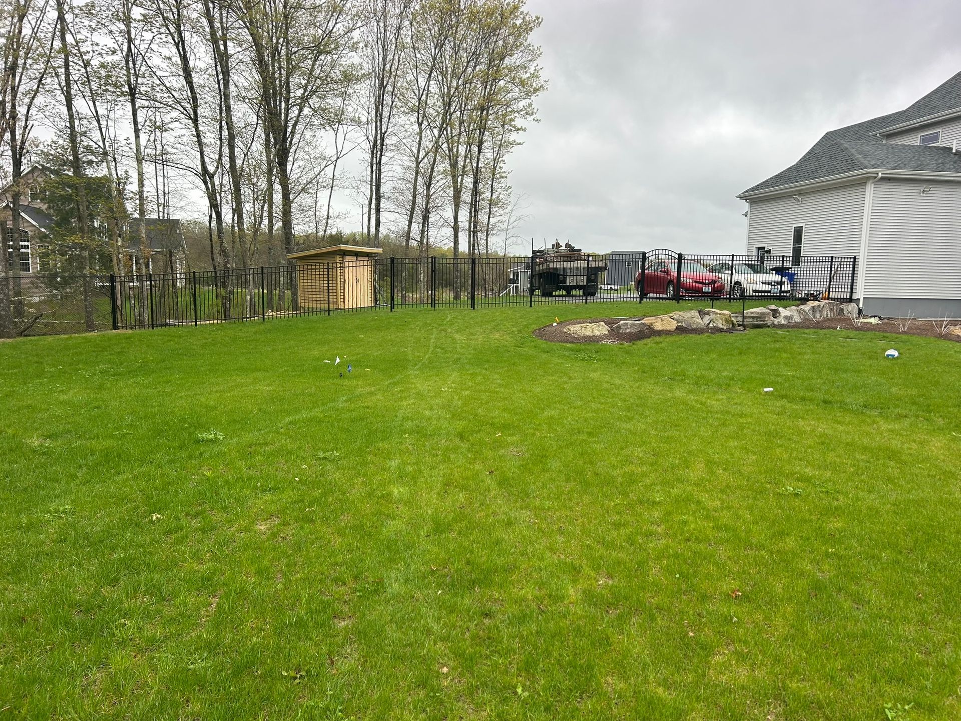 Backyard with green lawn, tall trees, a shed, and a house under cloudy skies.