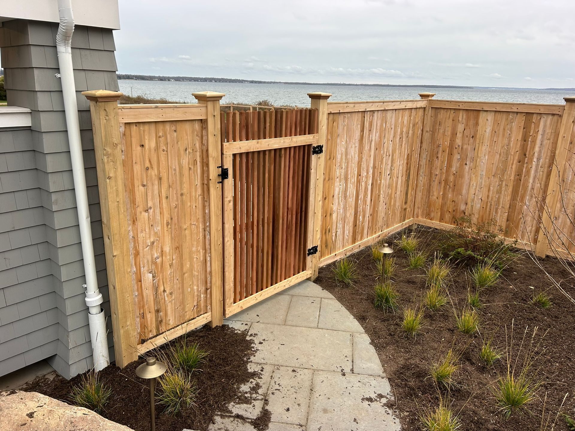 Wooden backyard fence gate beside a paved path, with the ocean visible beyond.