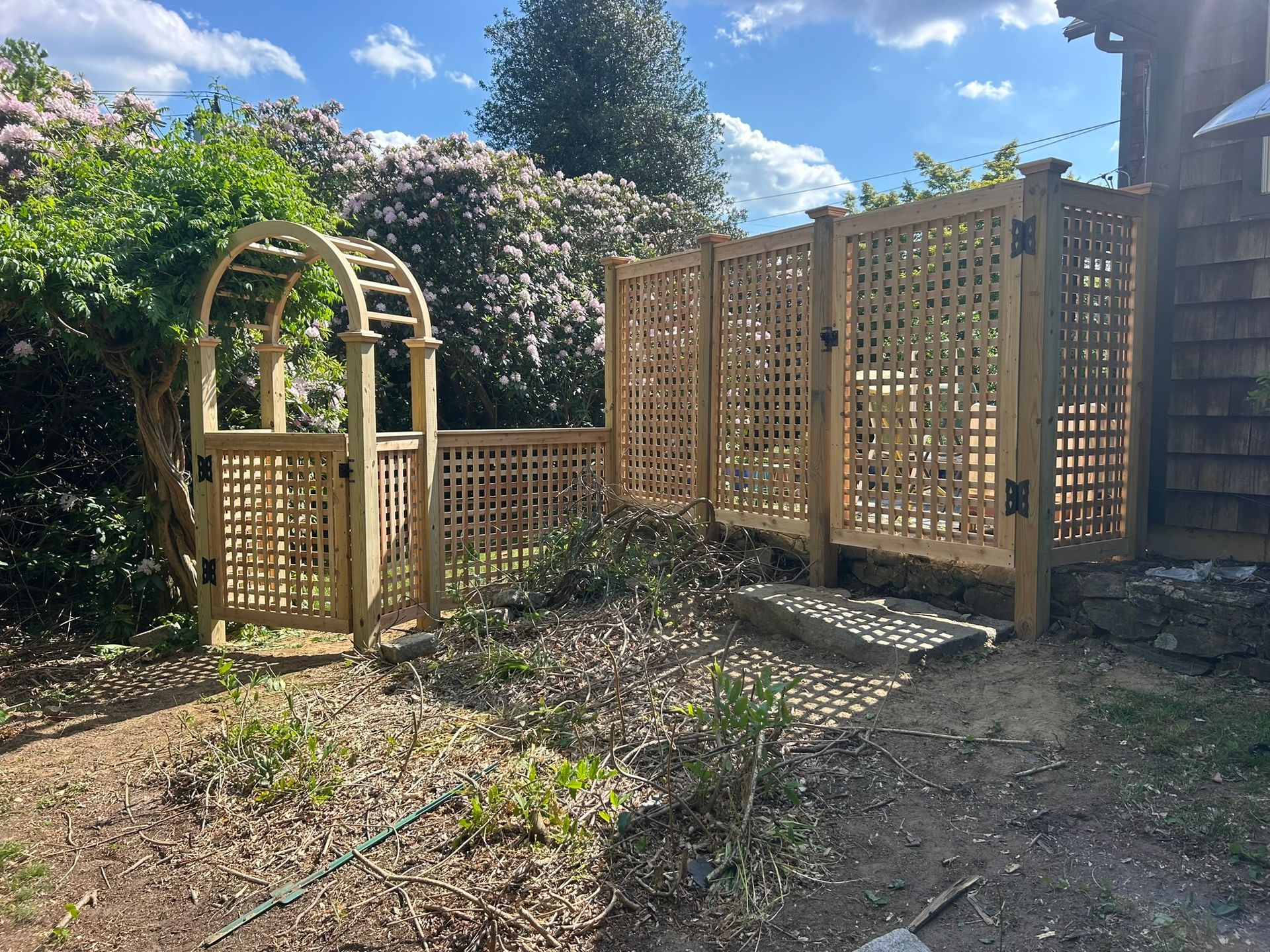 Wooden garden trellis and fenced patio beside a house, with a small arched gate in a sunny yard.