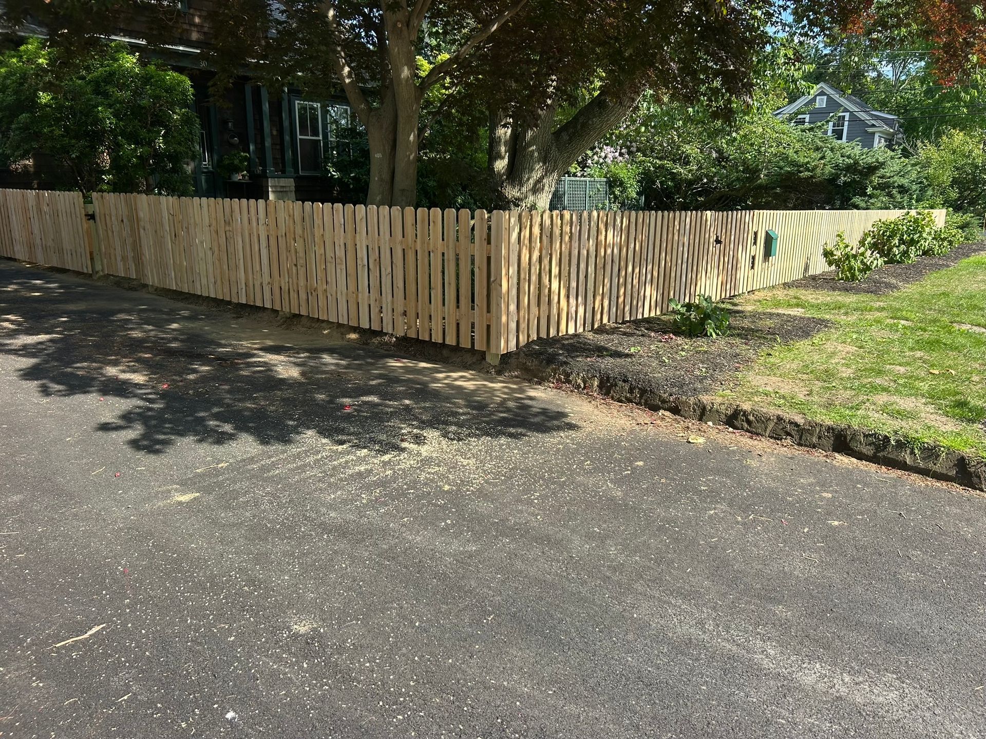 Light wooden picket fence along a roadside corner under trees, beside a grassy yard