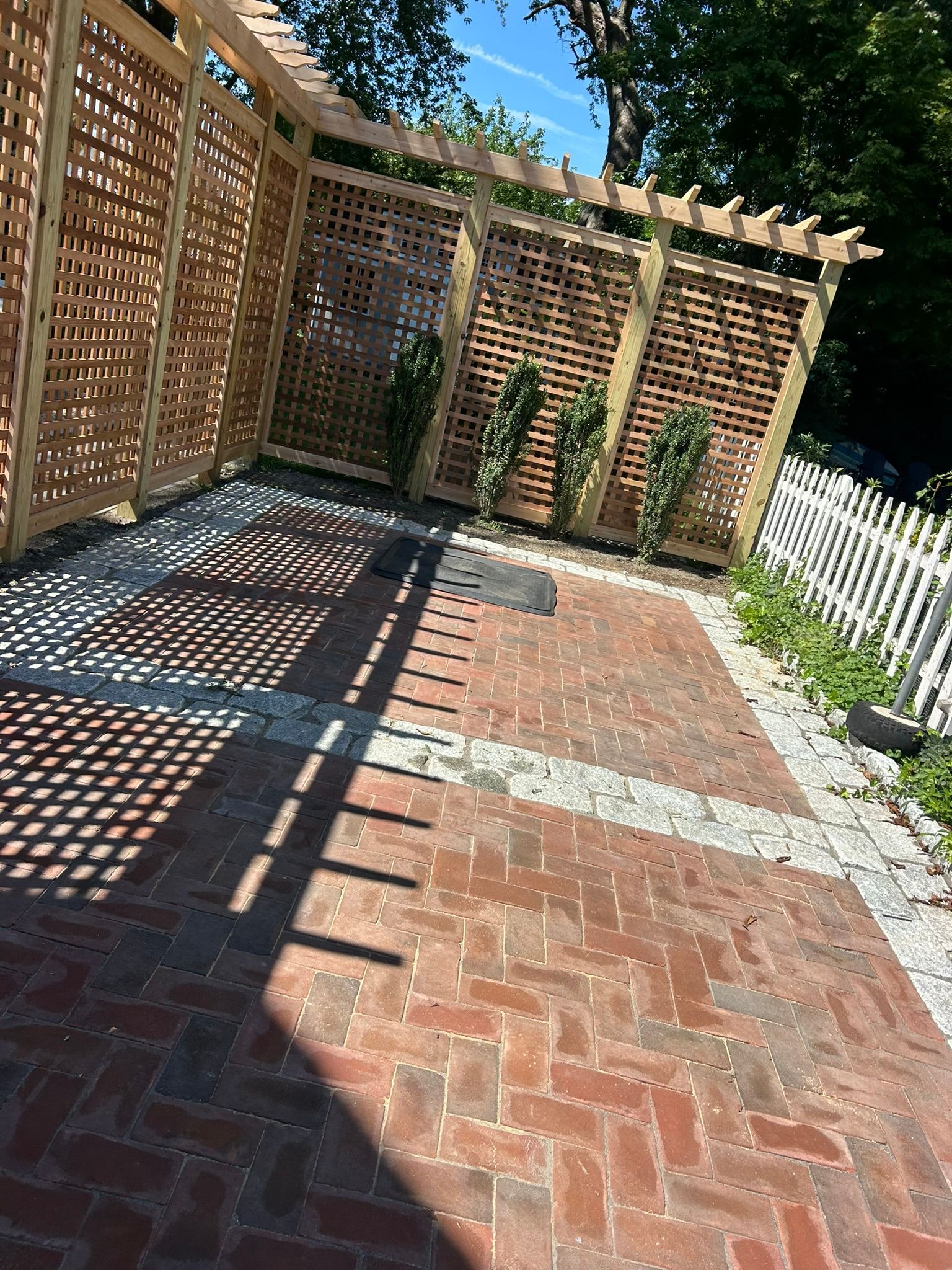 Brick patio with lattice wooden screens, potted shrubs, and strong sunlight casting shadows