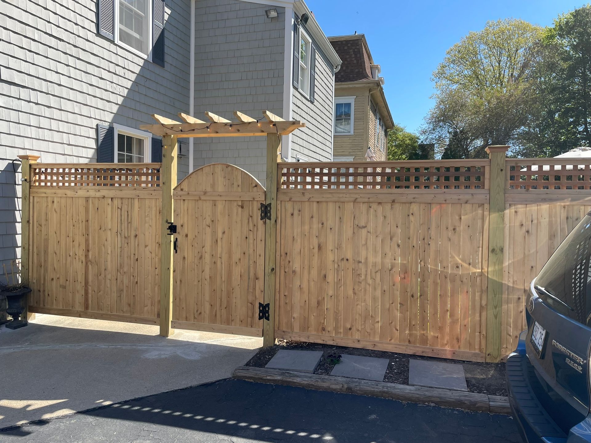 Wooden privacy fence and gate between two gray houses beside a driveway under a clear sky