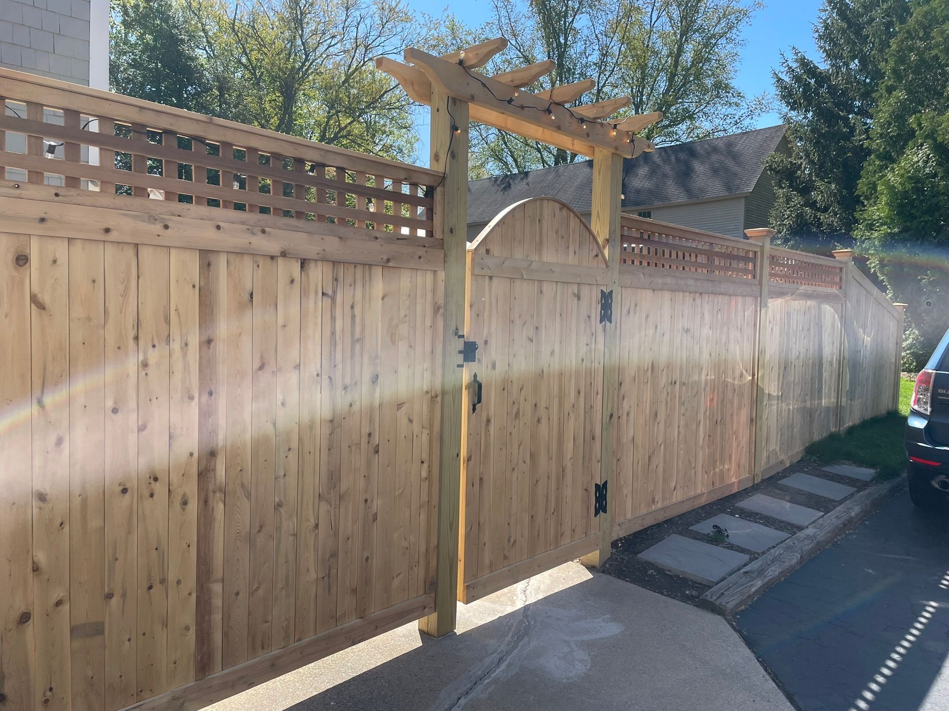 Wooden fence and gate beside a driveway, with a sunlit pergola and trees overhead.