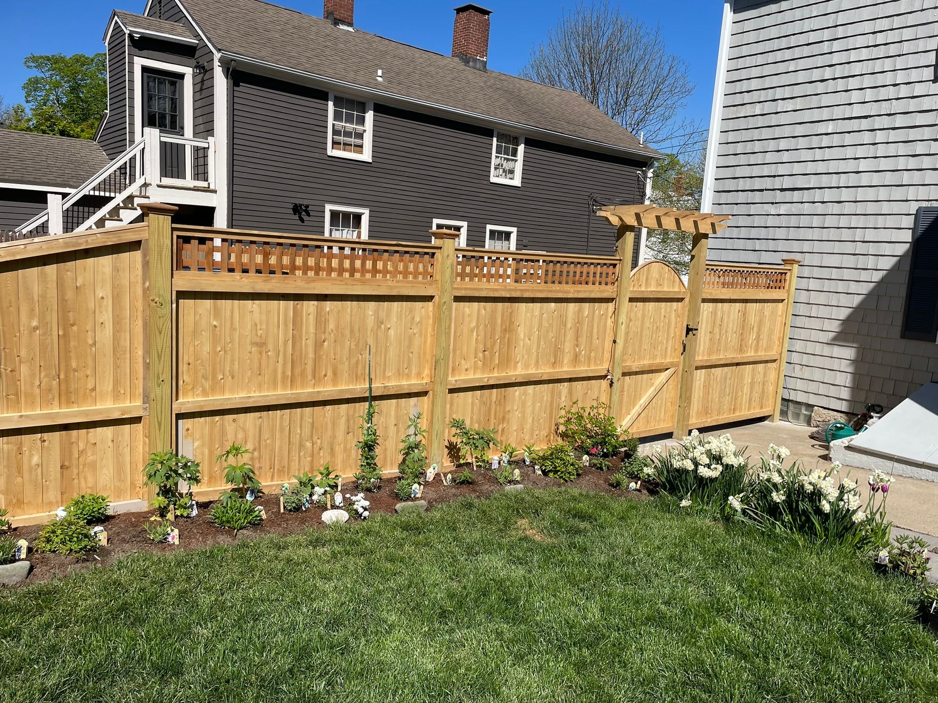 Backyard with a wooden fence, flower bed, and green lawn beside houses on a sunny day